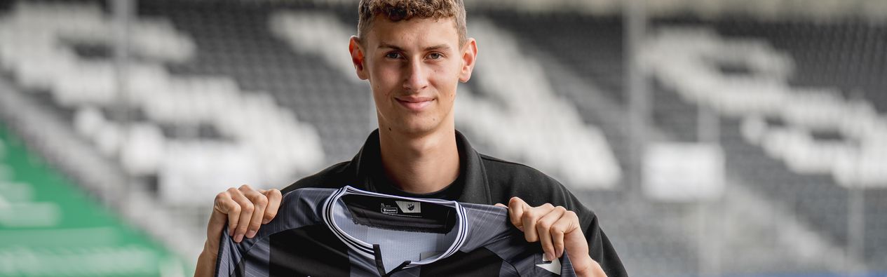 Benedikt Wimmer steht im Stadion und hält das Auswärtstrikot der Saison 2025/26 vor der Brust 